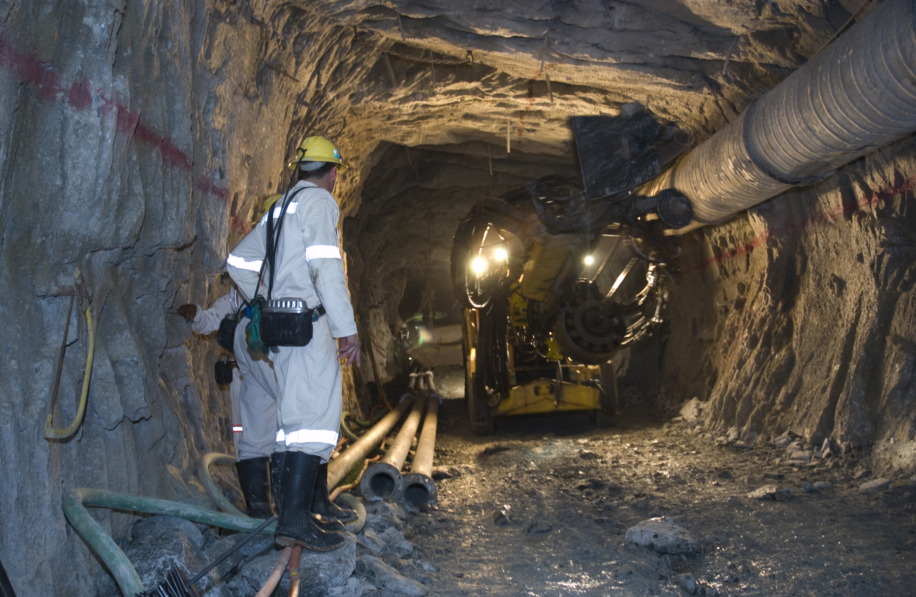 Homem parado no canto esquerdo da imagem, com roupa cinza com material refletor. Máquina utilizada para perfurar rochas no canto direito. Tudo em uma mina subterrânea.