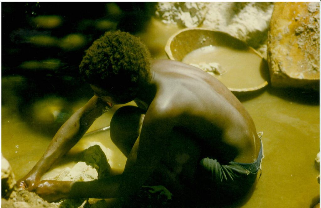 Rapaz negro à esquerda da foto, abaixado, como se estivesse lavando um pano no rio. A água do rio é amarelada, devido ao processo de garimpo de diamante.