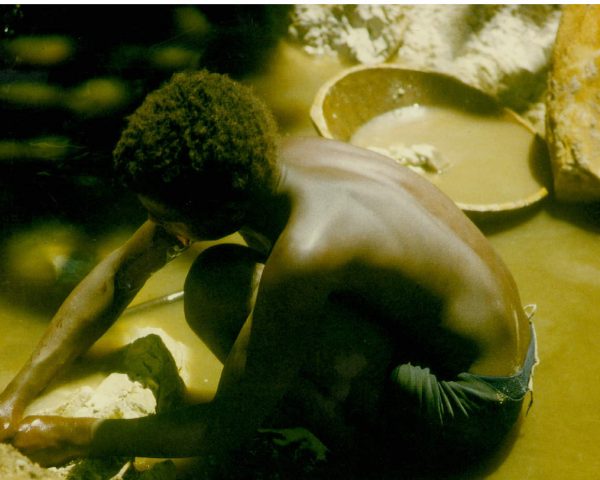 Rapaz negro à esquerda da foto, abaixado, como se estivesse lavando um pano no rio. A água do rio é amarelada, devido ao processo de garimpo de diamante.