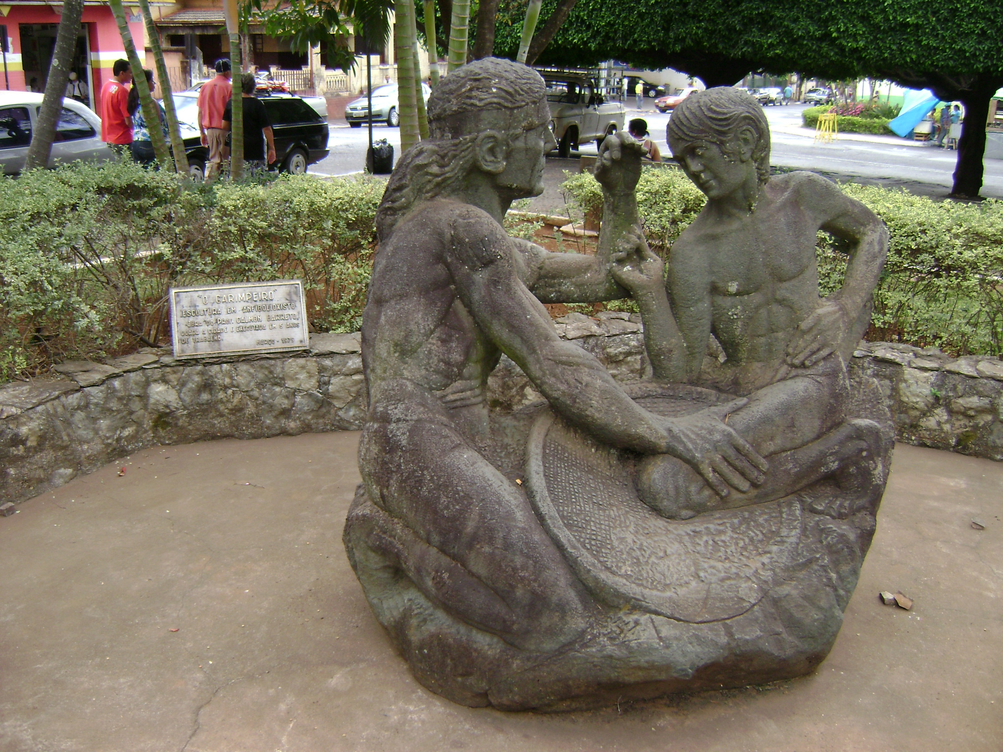 Escultura de dois homens debruçados sobre uma peneira, observando uma pedra que um deles segura. 